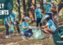 A group of volunteers wearing protective gloves, carrying trash bags, and picking up discarded plastic litter in a nature preserve.